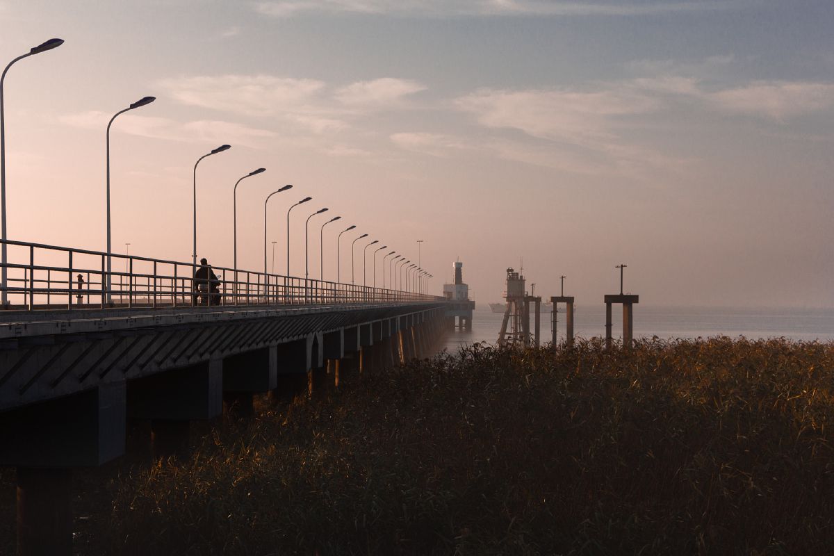 A long pier stretches into calm waters at dusk, lined with street lamps leading toward distant structures.