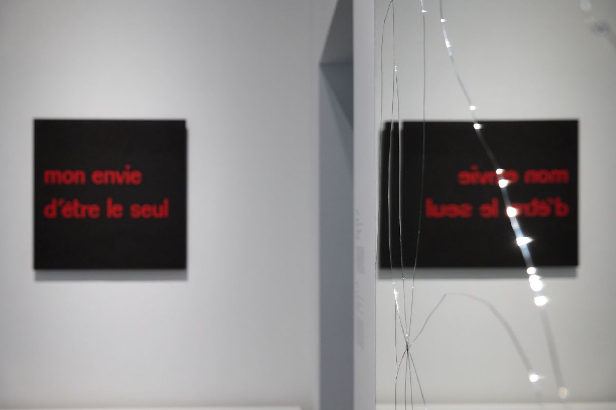 Glowing red French text on black panels reflects in a mirror, intersected by delicate strands of light in a minimalist gallery space.
