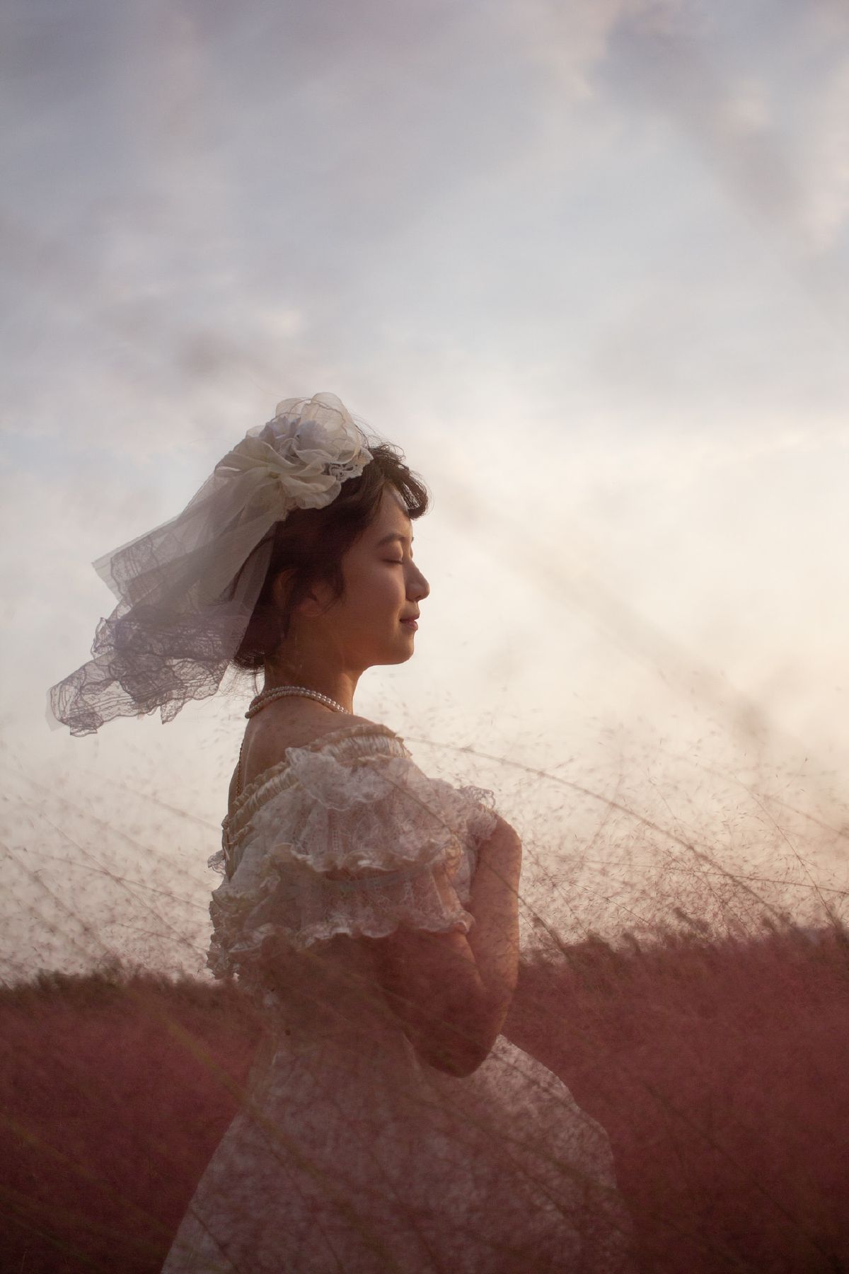 A bride in vintage lace and pearls stands serenely among swaying pink grasses as golden sunset light bathes the scene.