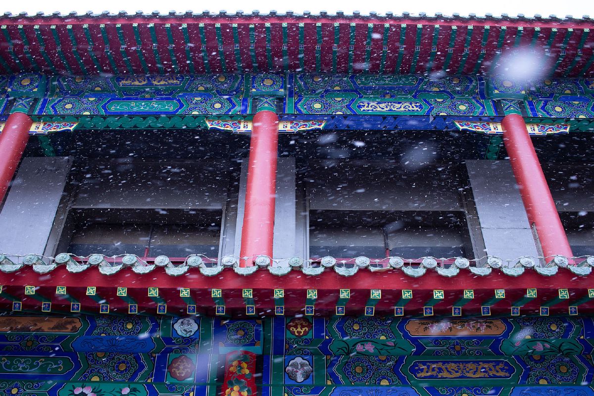 Snow falls gently on an ornate traditional Chinese building with vibrant painted eaves and crimson pillars.