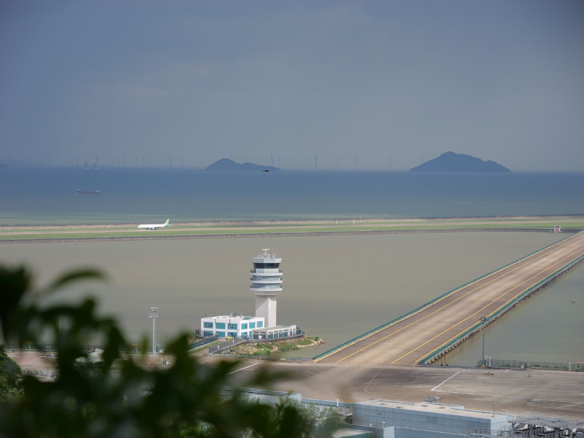 An airport control tower stands sentinel beside a runway that stretches across reclaimed land toward distant mountains and wind turbines on the horizon.