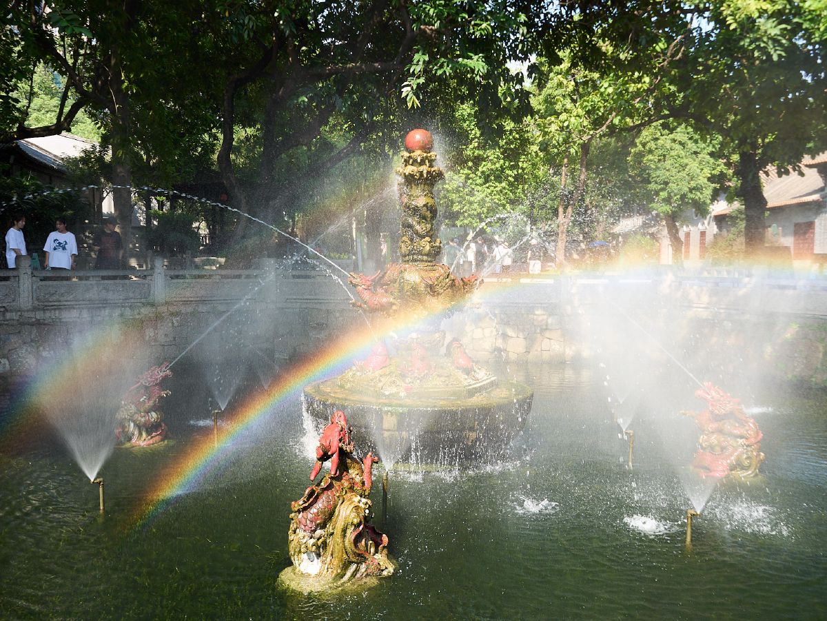 A vibrant rainbow arcs across a traditional Chinese fountain where dragon statues spout water into a serene pond.