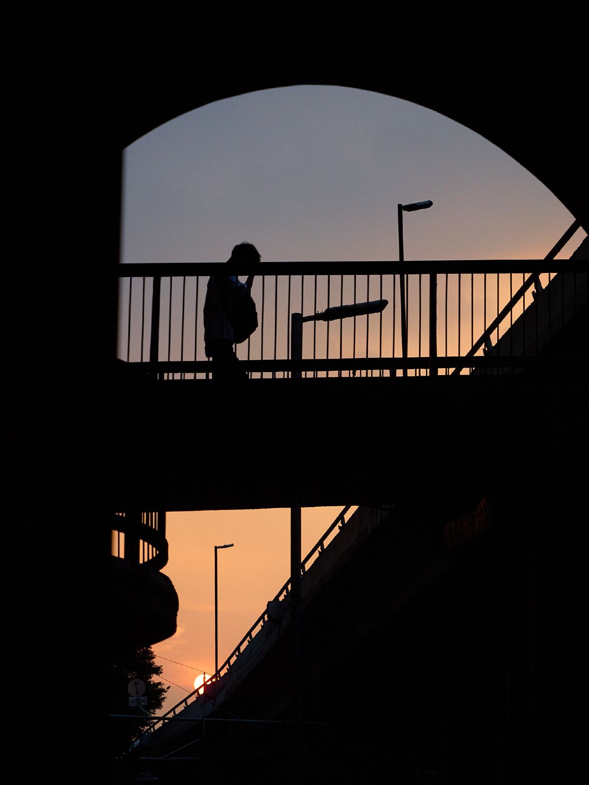A lone pedestrian is silhouetted against the warm glow of sunset while crossing an arched footbridge.