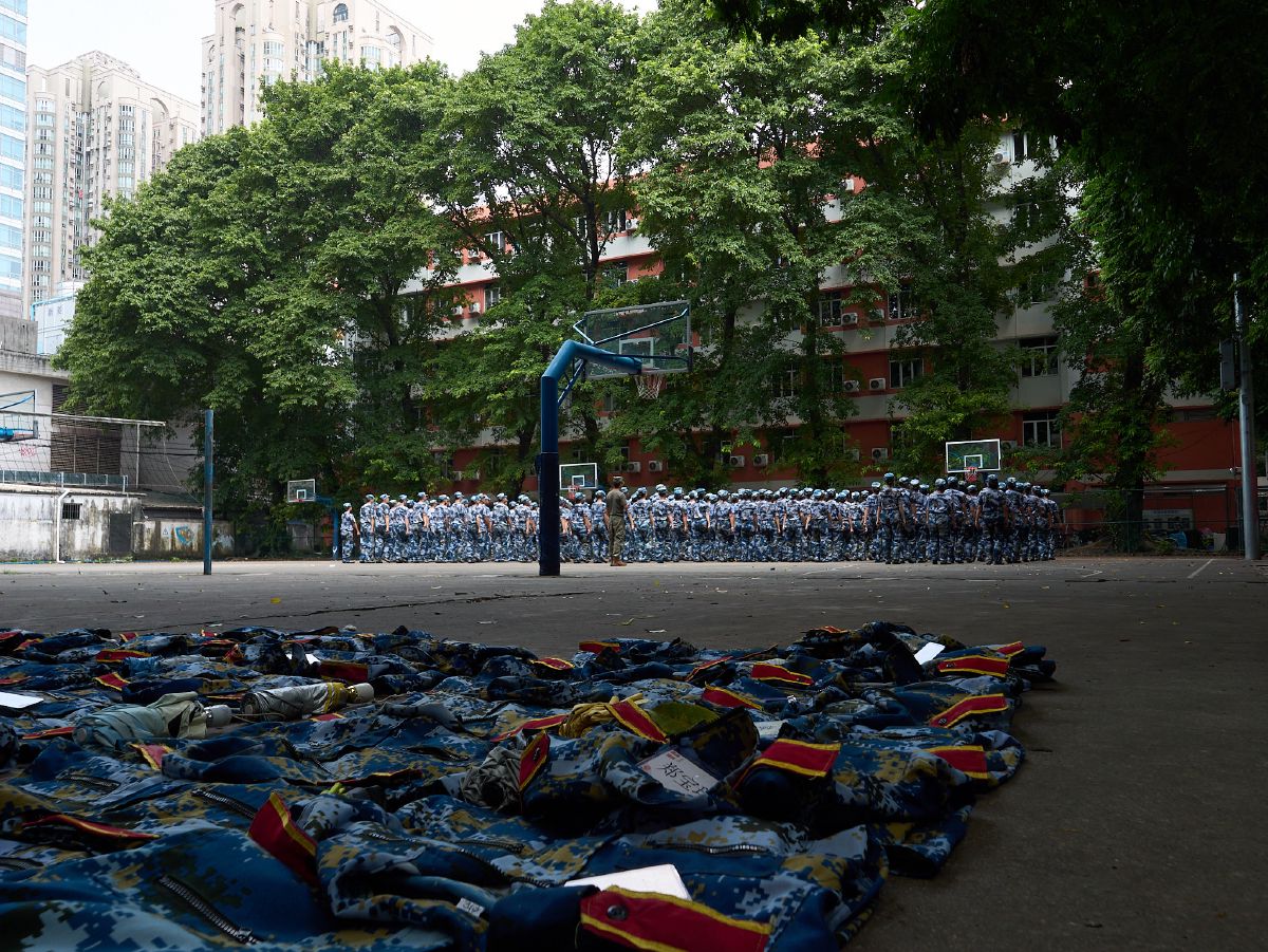 Rows of folded camouflage uniforms await students standing in formation during military training on a school basketball court.