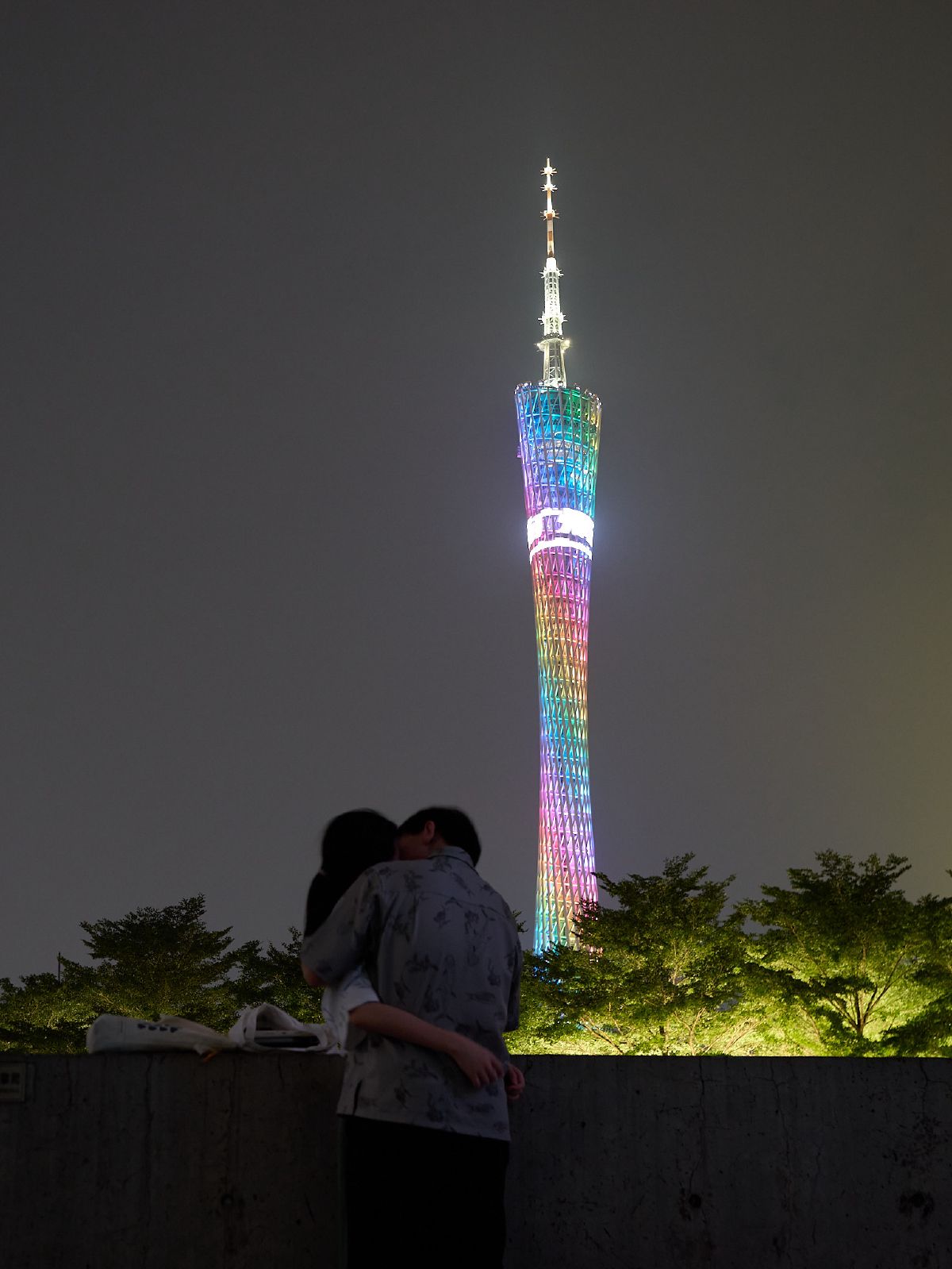 A couple embraces on a ledge overlooking the illuminated Canton Tower glowing with rainbow lights against the night sky.
