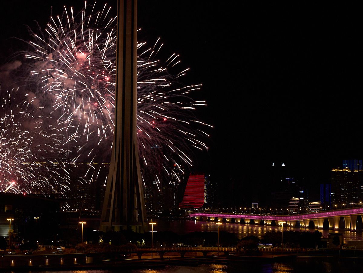 Brilliant white fireworks burst above the Macau Tower while a pink-lit bridge spans the dark waters below.