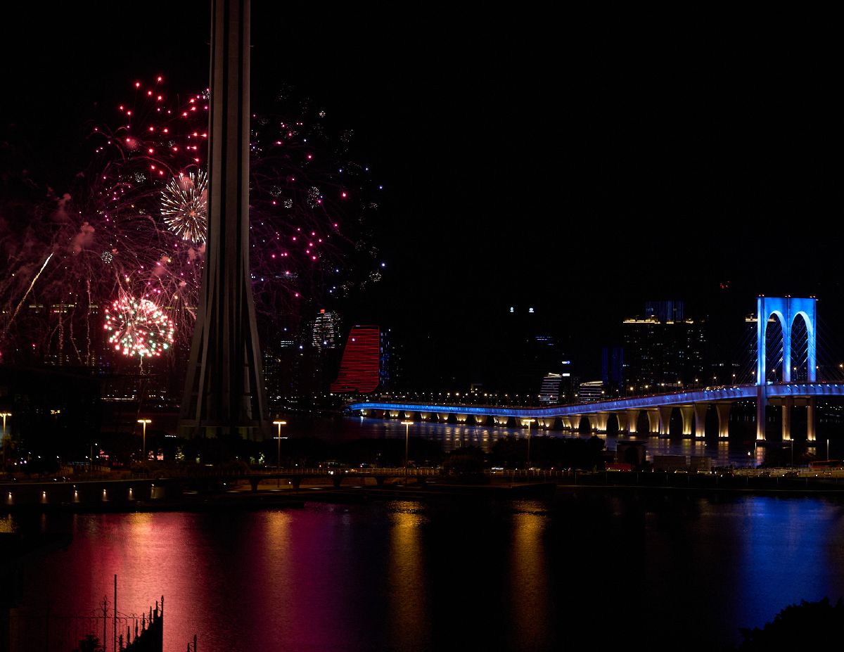 Red and gold fireworks illuminate the night sky near the Macau Tower while colorful reflections dance on the water's surface.