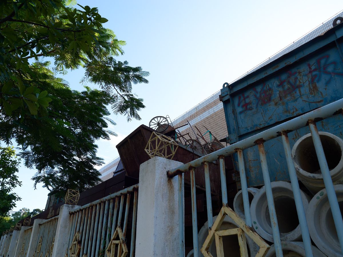 Concrete pipes and rusted metal containers sit behind a weathered fence partially shaded by overhanging tree branches.