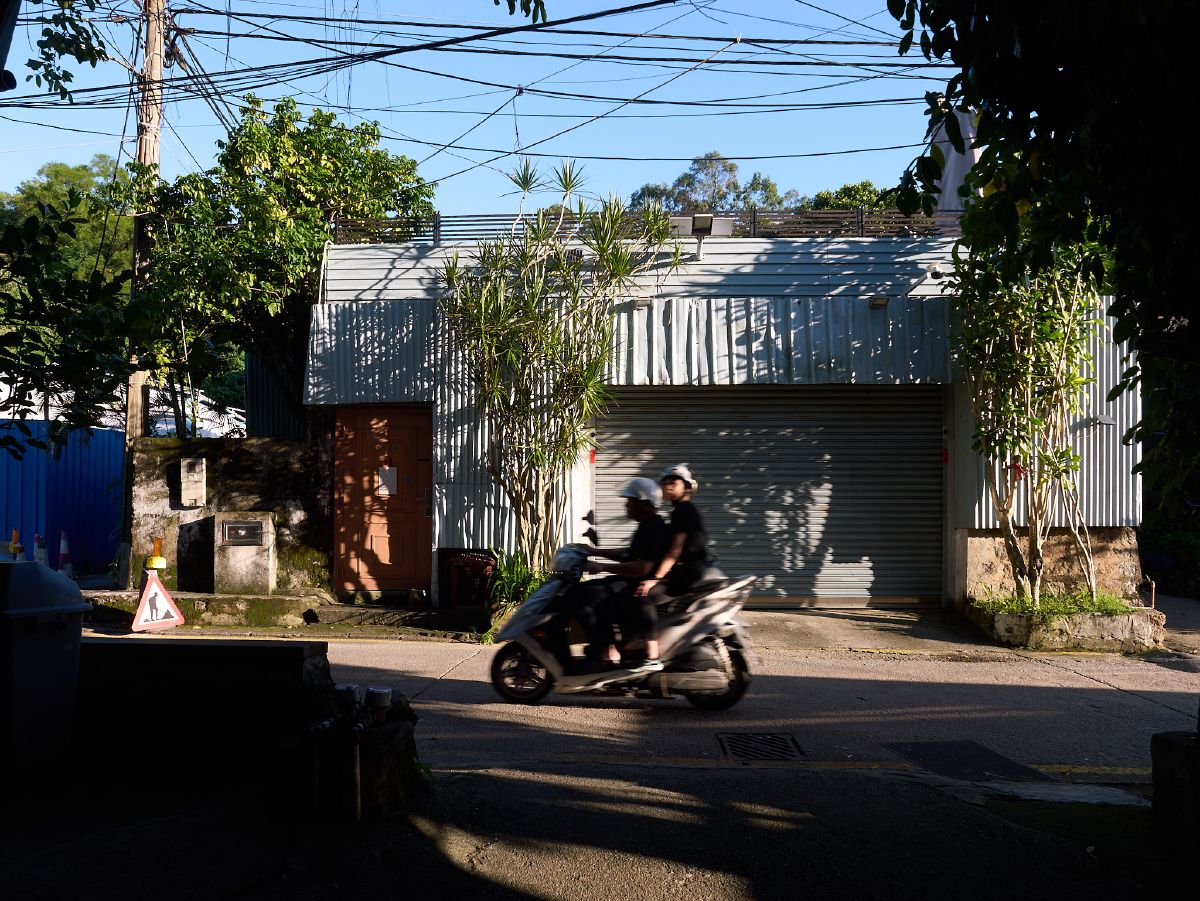 Two helmeted riders on a scooter cruise past a corrugated metal warehouse bathed in dappled afternoon sunlight.