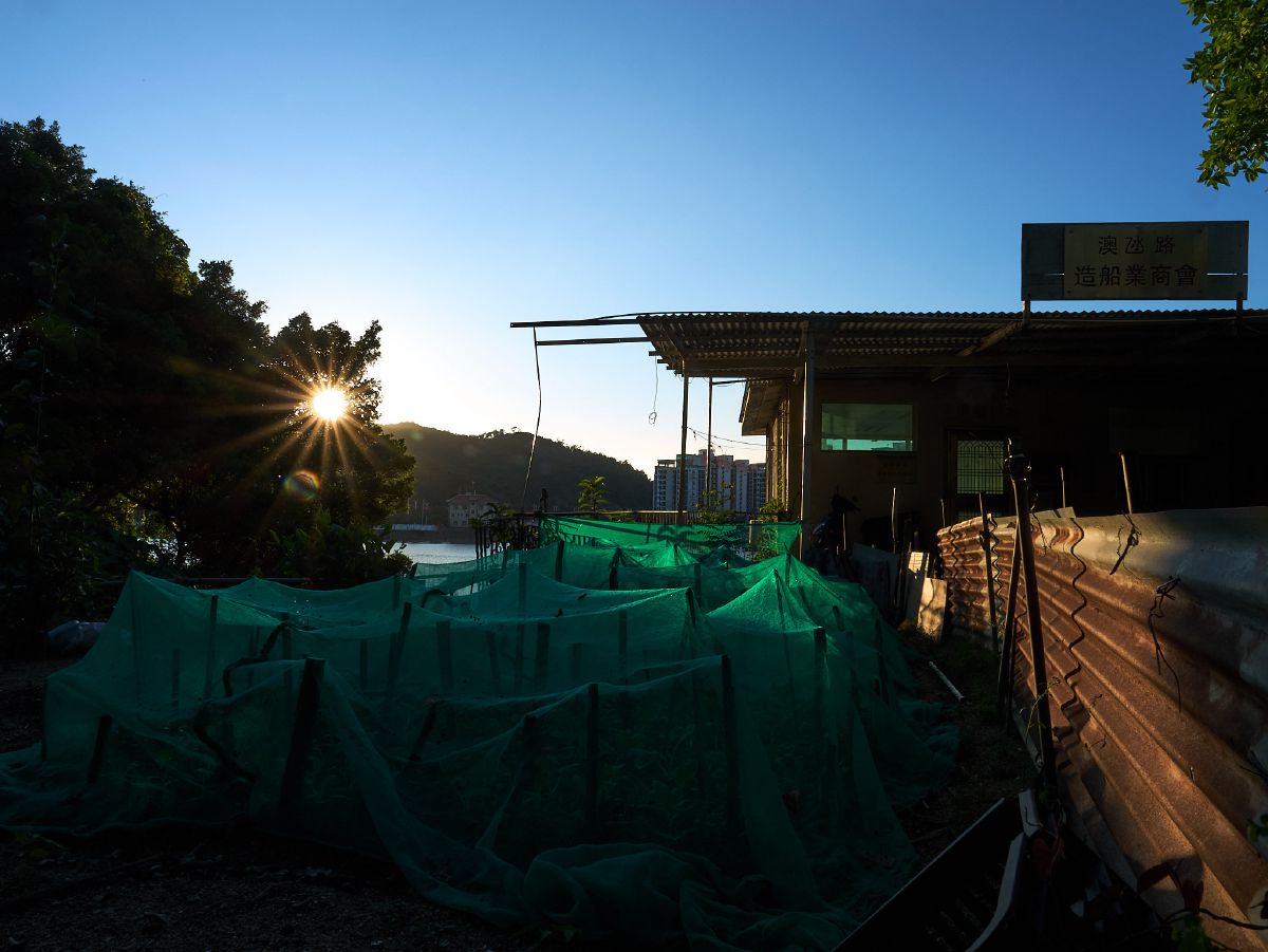 The setting sun casts golden rays across green fishing nets drying near a small harbor building in Coloane.