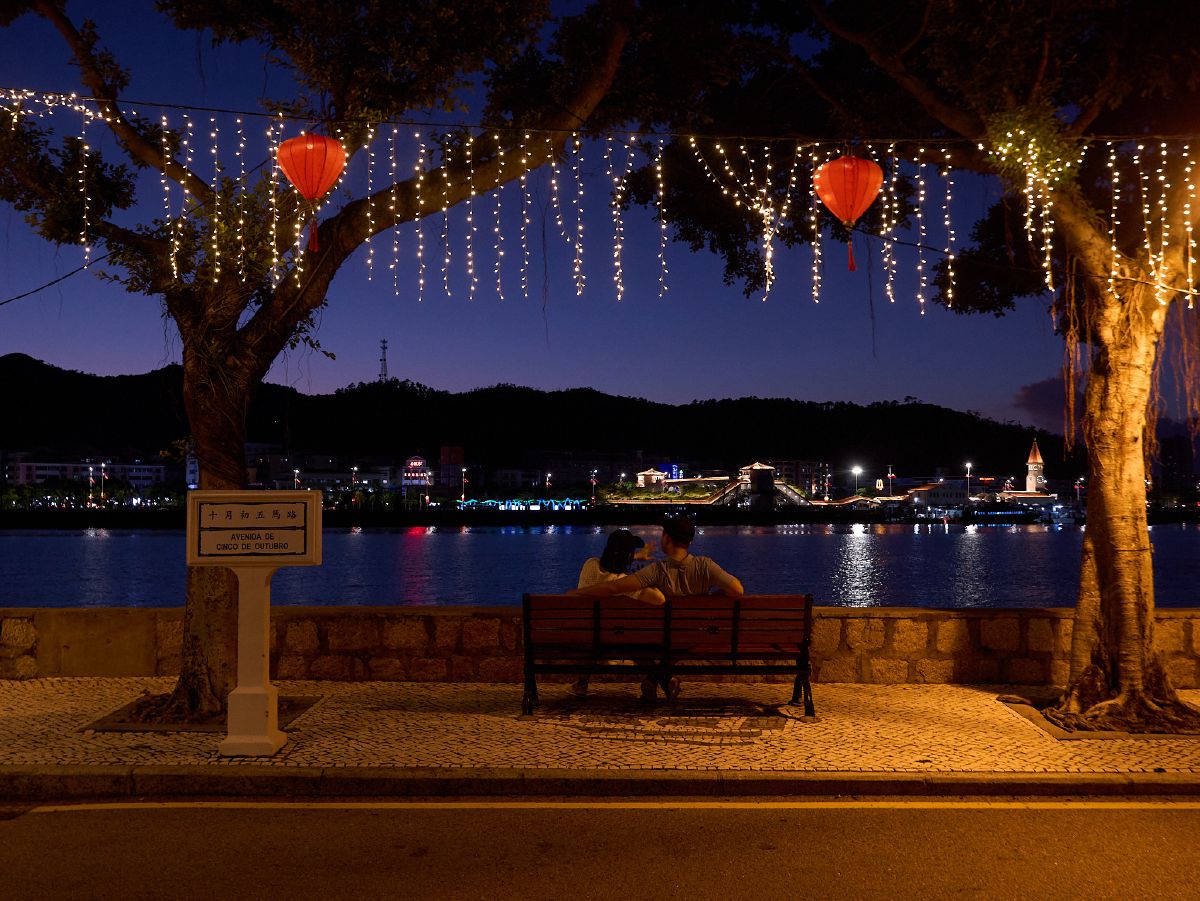 A couple sits on a bench beneath trees adorned with twinkling lights and red lanterns, overlooking the tranquil waters of Coloane at dusk.