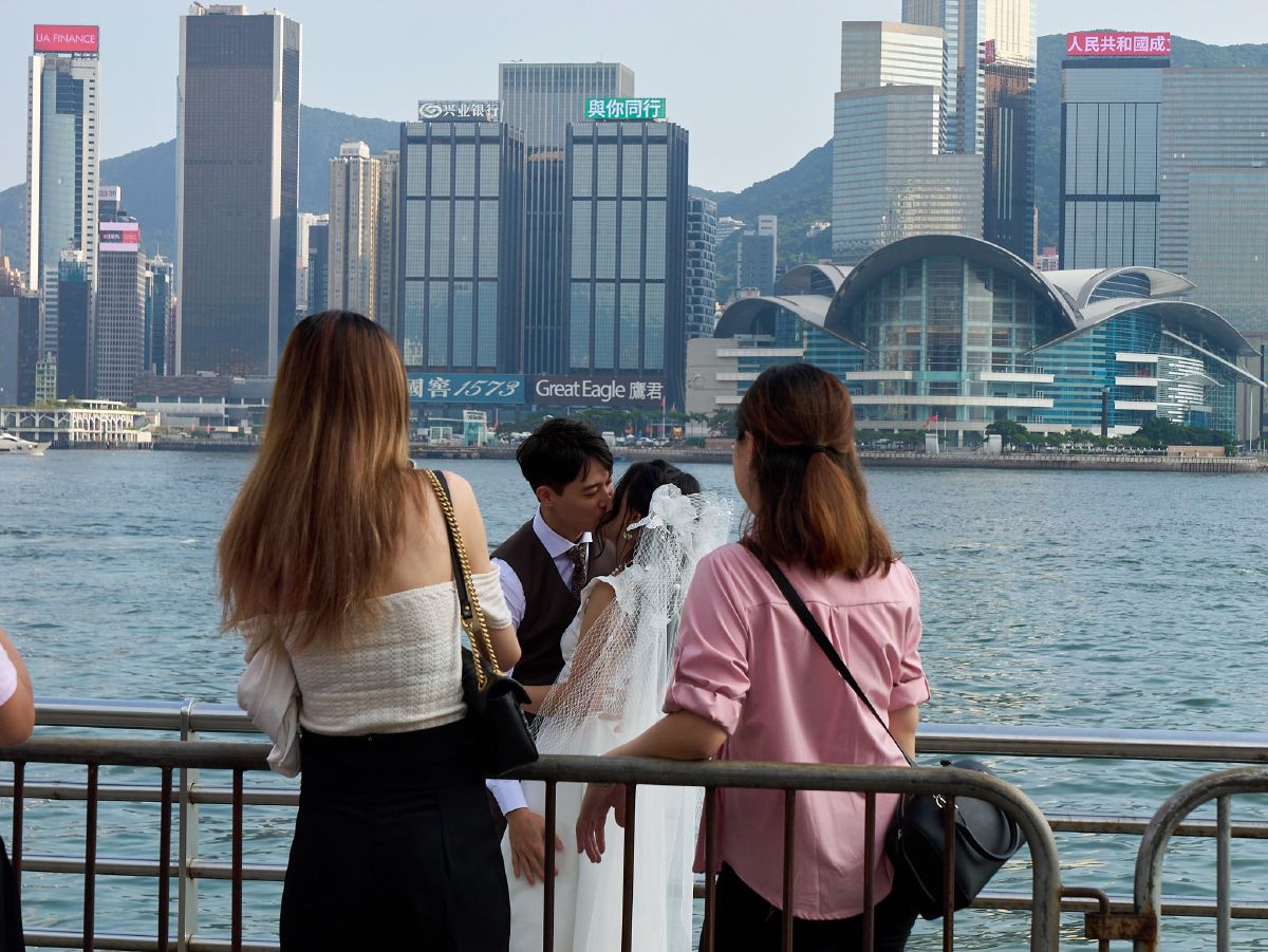A newlywed couple shares a kiss on the waterfront promenade with Hong Kong's iconic skyline and convention center as their backdrop.