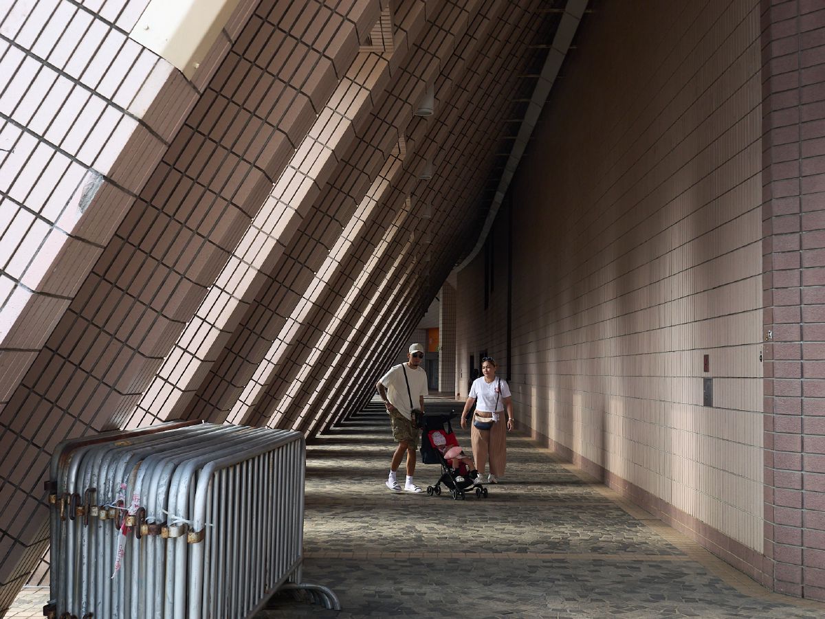 A family with a stroller walks through a dramatic corridor of angled tiled walls casting striped shadows across the pathway.