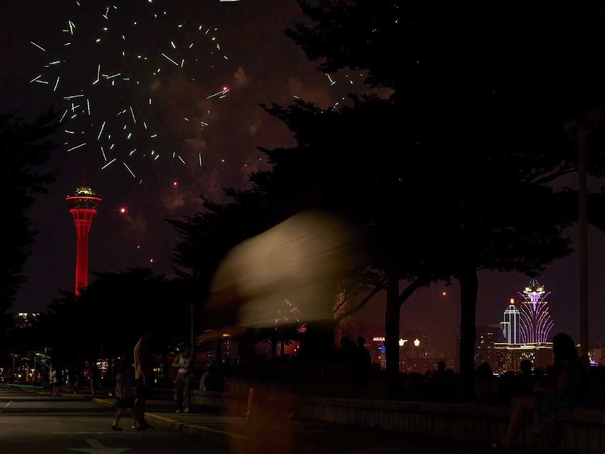 Spectators watch green and gold fireworks burst over the Macau skyline, with the iconic red tower illuminated against the night sky.