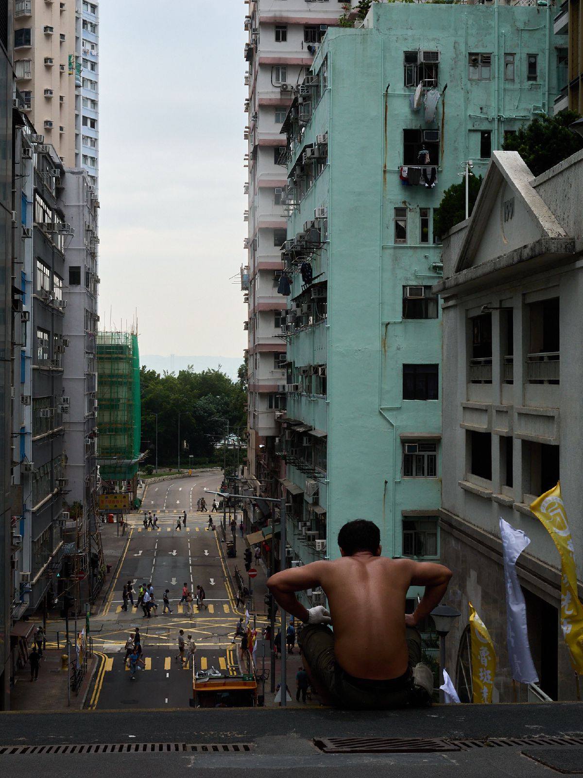 A shirtless worker pauses on a steep street overlooking a canyon of pastel apartment towers leading down to the sea.
