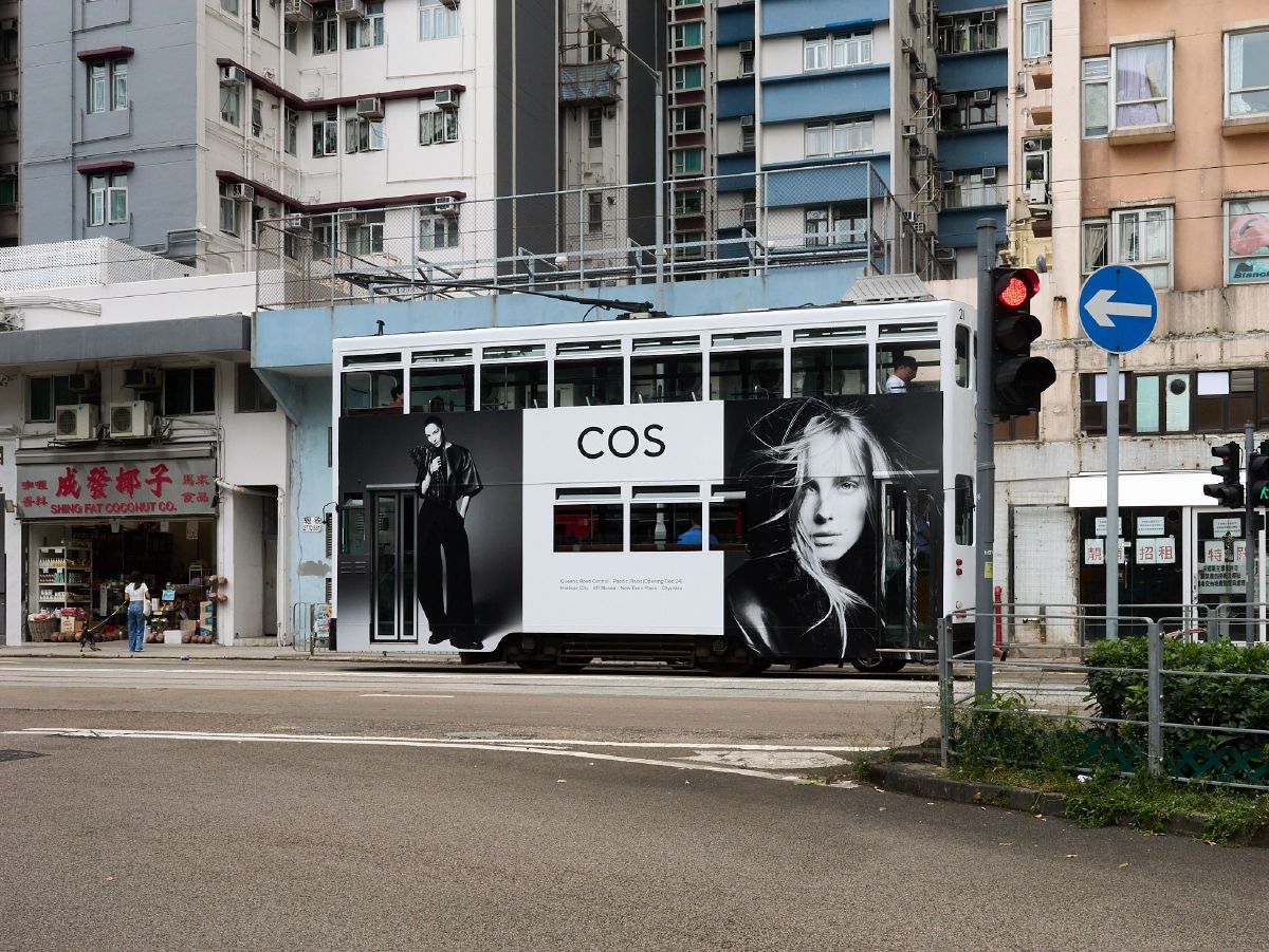 A classic double-decker tram wrapped in a COS fashion advertisement travels past colorful residential buildings in Sai Ying Pun.