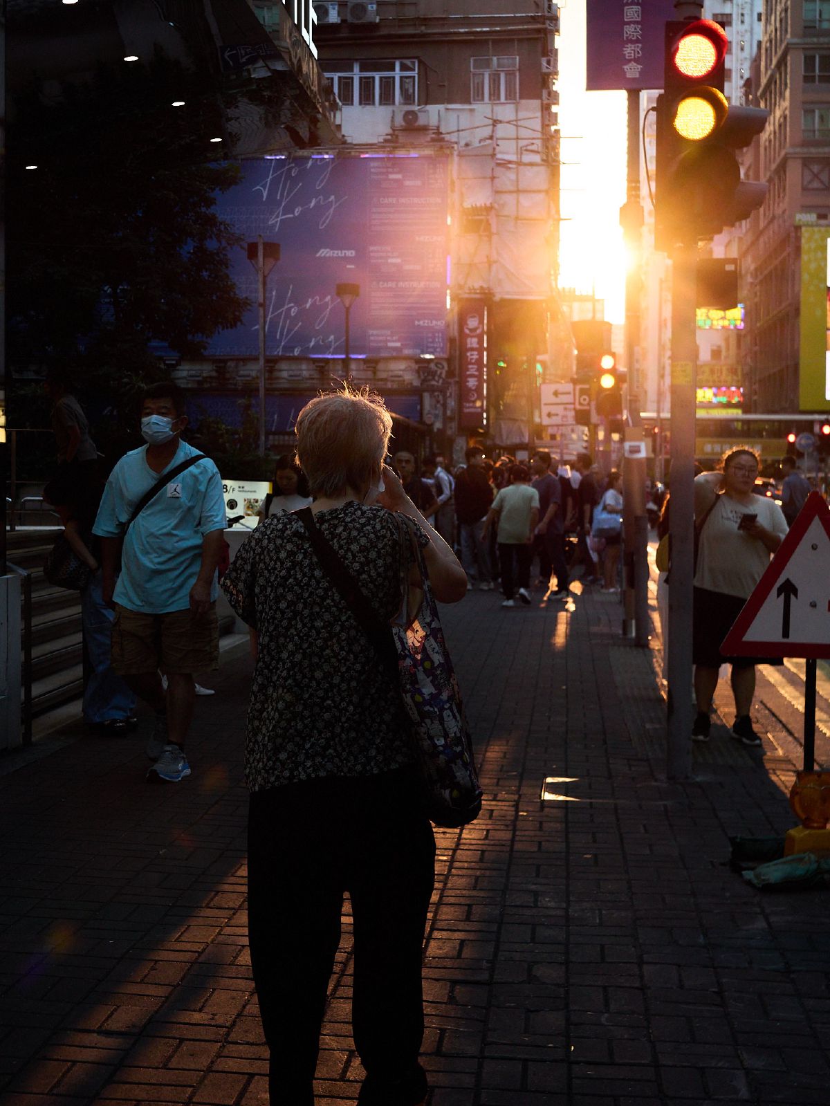 An elderly woman walks toward the golden sunset streaming between tall buildings on a busy Hong Kong sidewalk.