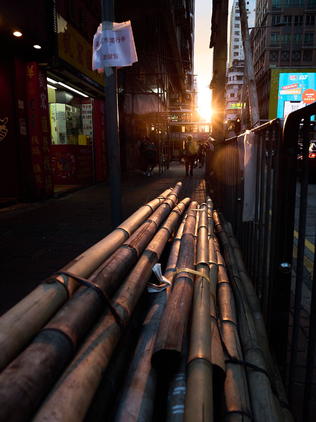 Bamboo scaffolding poles lie stacked along a narrow alleyway as the setting sun casts long shadows toward the distant harbor.