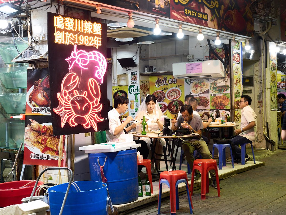 Diners enjoy a late-night meal at an outdoor seafood restaurant illuminated by a bright pink neon crab sign in Hong Kong.
