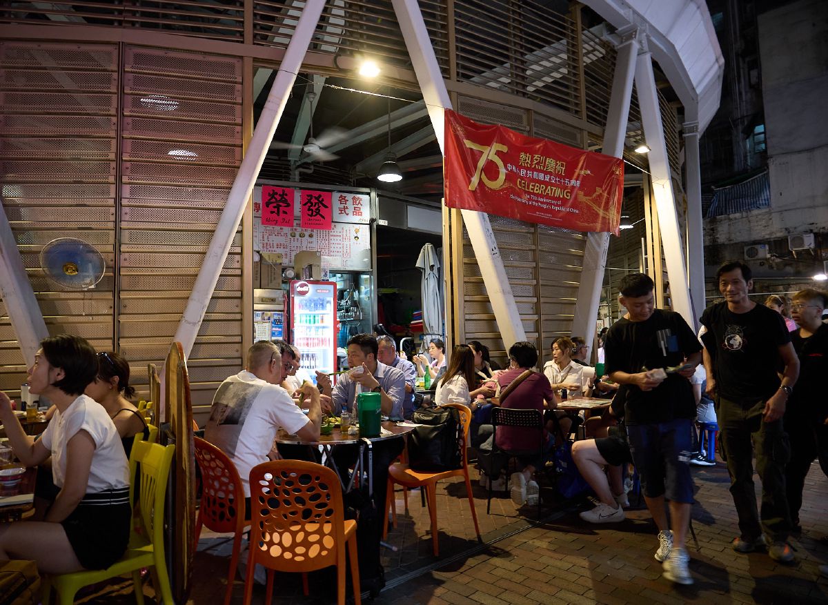 Patrons gather at tables outside a bustling nighttime eatery beneath a pedestrian bridge in an urban Hong Kong neighborhood.