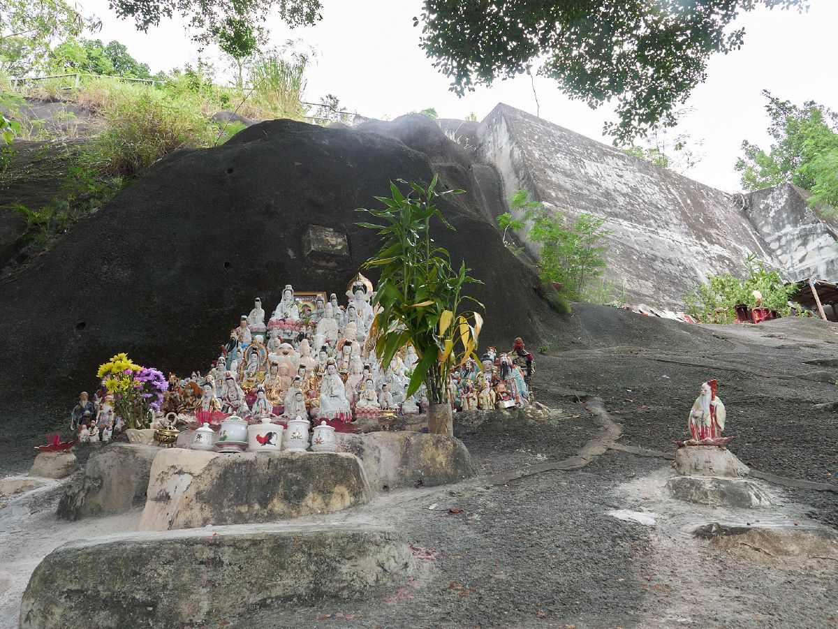 Numerous small Buddha and deity statues arranged on stone platforms against a rocky hillside create a peaceful shrine setting.