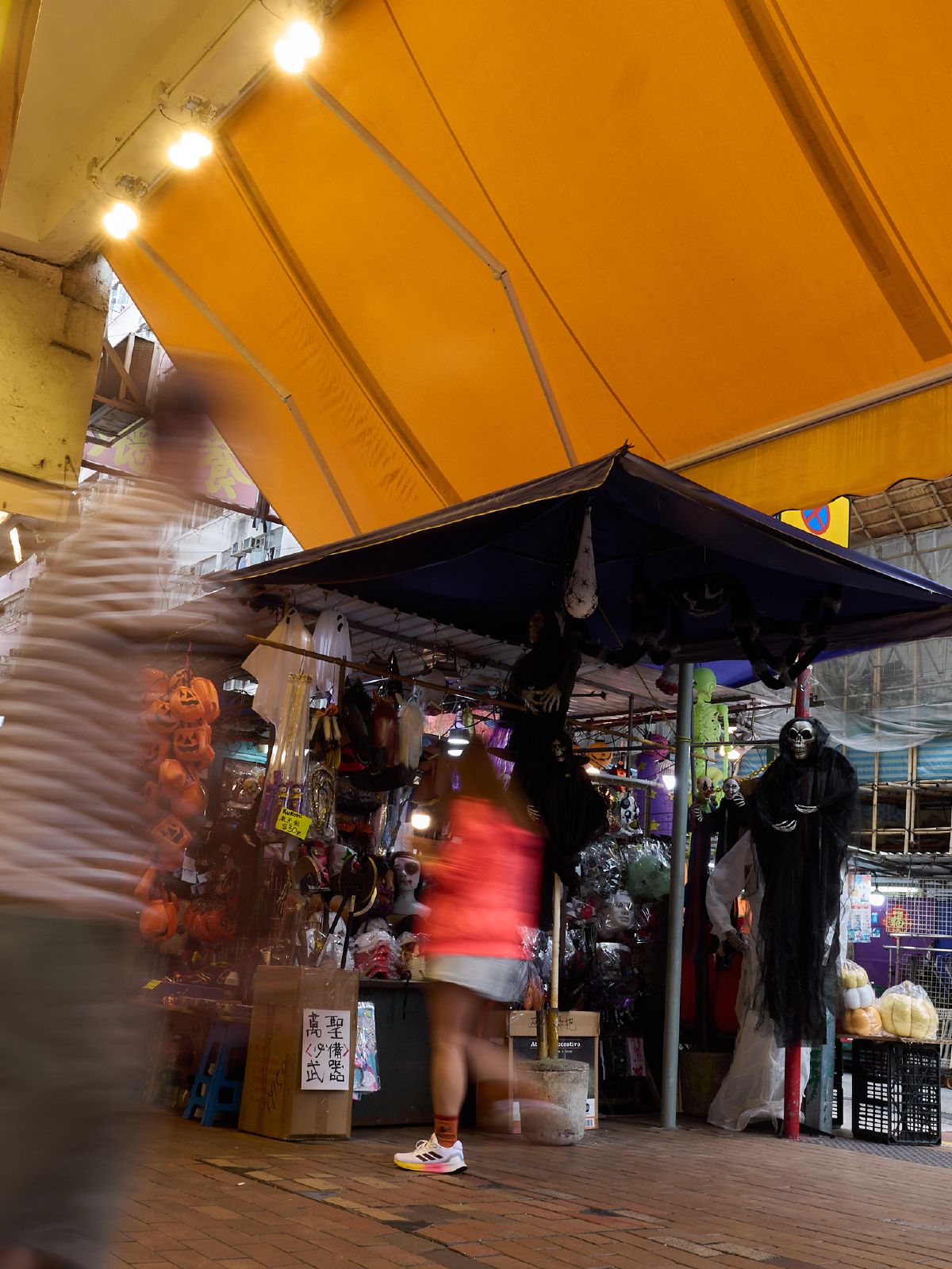 A street vendor stall selling Halloween decorations and costumes draws passersby under a bright yellow overhead structure.