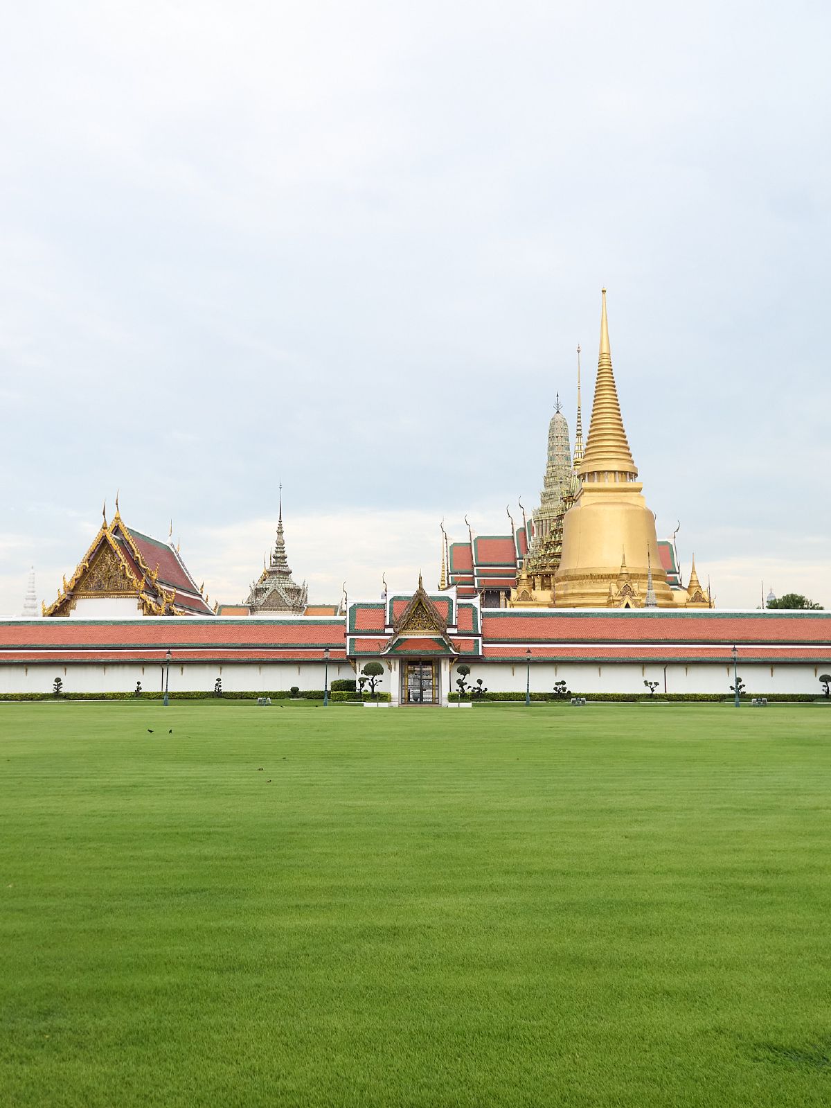 The golden spires and ornate rooftops of the Grand Palace rise above a manicured green lawn in Bangkok.