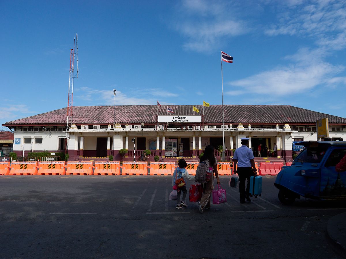 Travelers with luggage walk toward the entrance of Ayutthaya Railway Station under a bright blue sky.