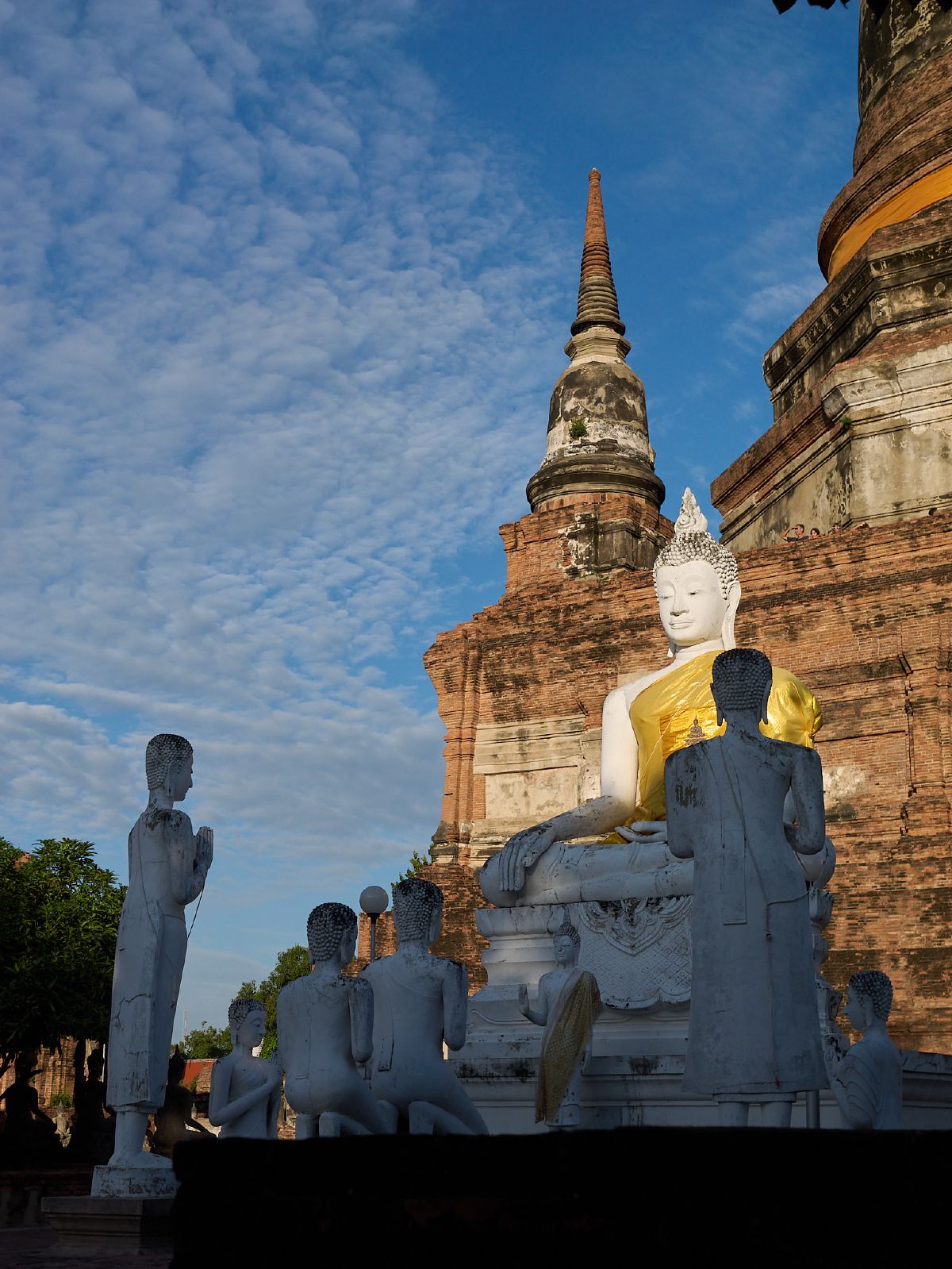 A white Buddha statue draped in golden cloth sits serenely among smaller disciple figures at an ancient Thai temple.