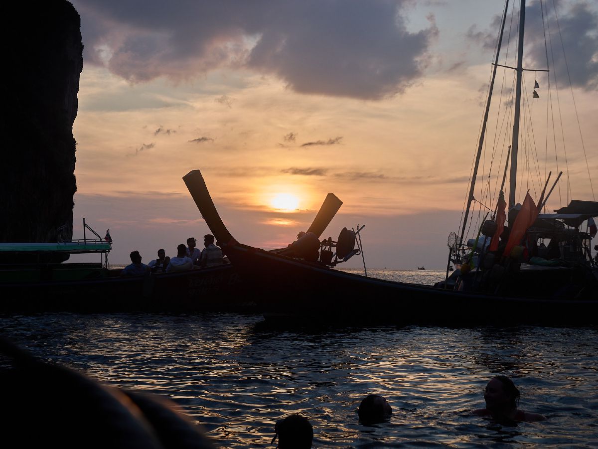 Longtail boats silhouetted against a golden Krabi sunset, with swimmers floating in the warm Andaman Sea as day fades to evening.