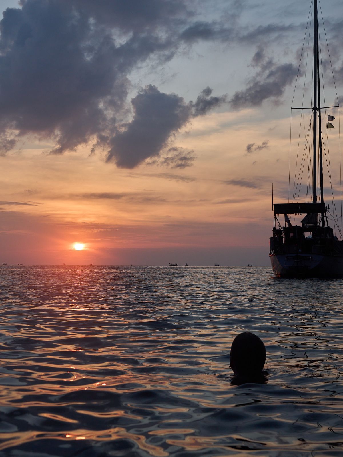 A lone swimmer watches the sun sink into the horizon off Krabi's coast, while a sailboat drifts peacefully on the amber-colored water.