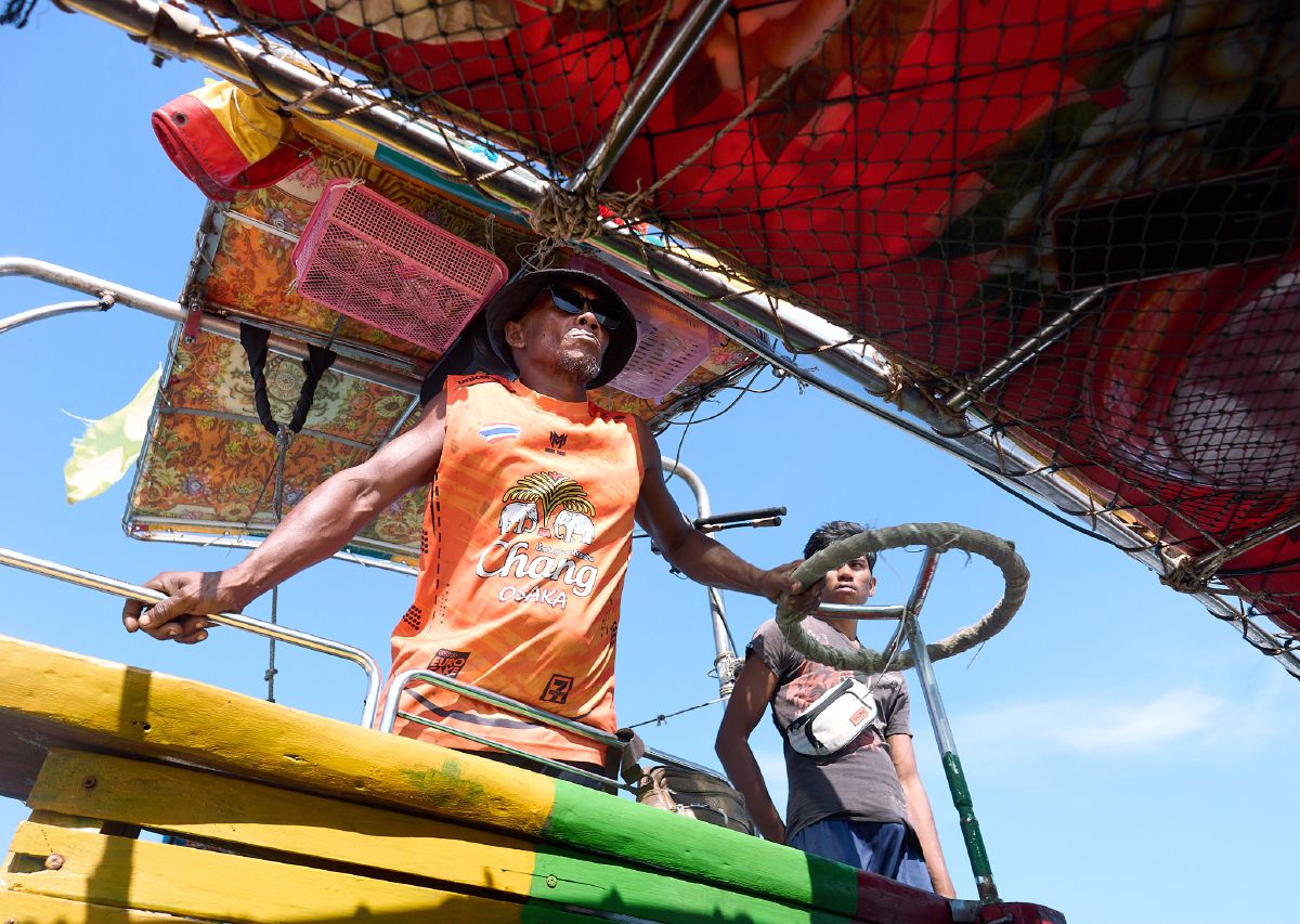 A boat captain steers his colorful vessel through Krabi's waters, navigating with a large rope-wrapped wheel under a bright blue sky.