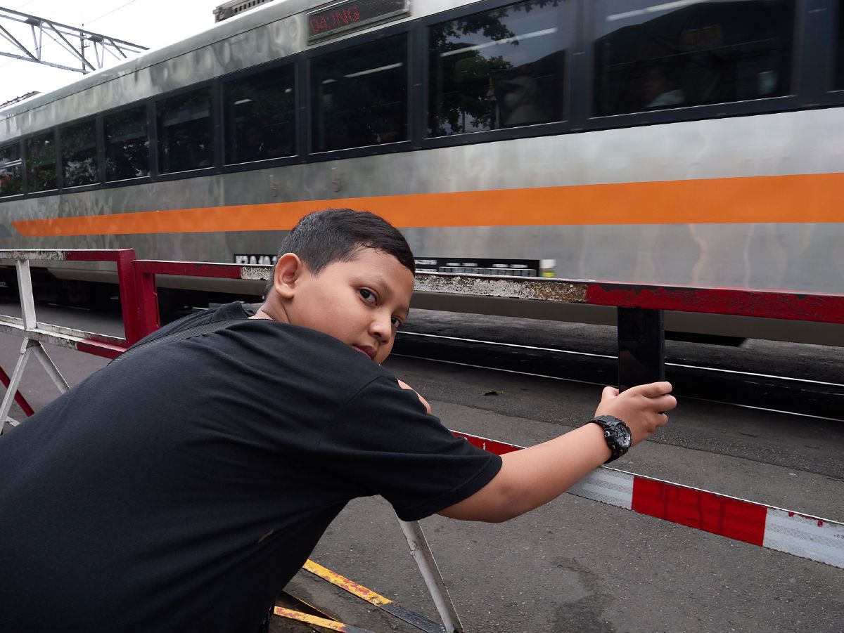 A young boy leans against a railway crossing barrier in Yogyakarta as a silver train passes behind him.