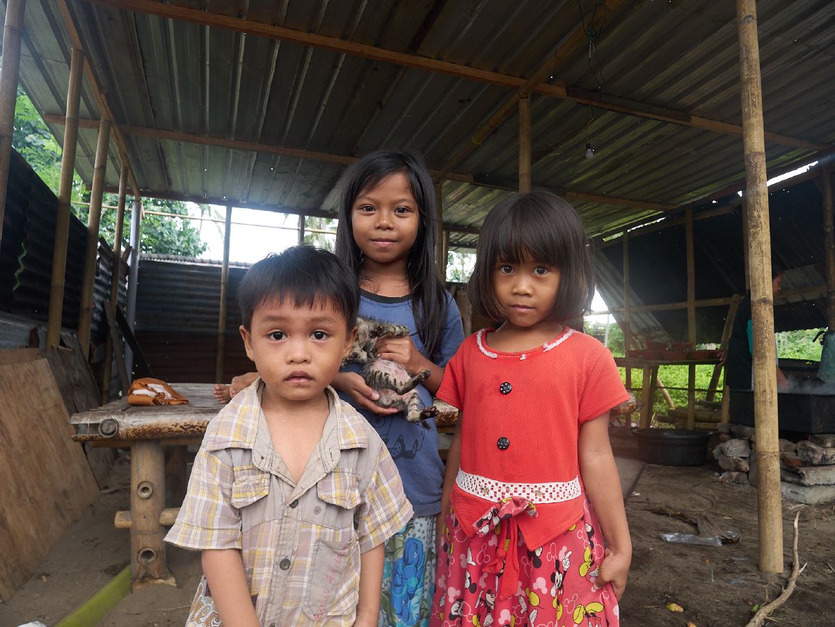 Three children stand together in a rustic wooden shelter, one gently holding a small kitten.