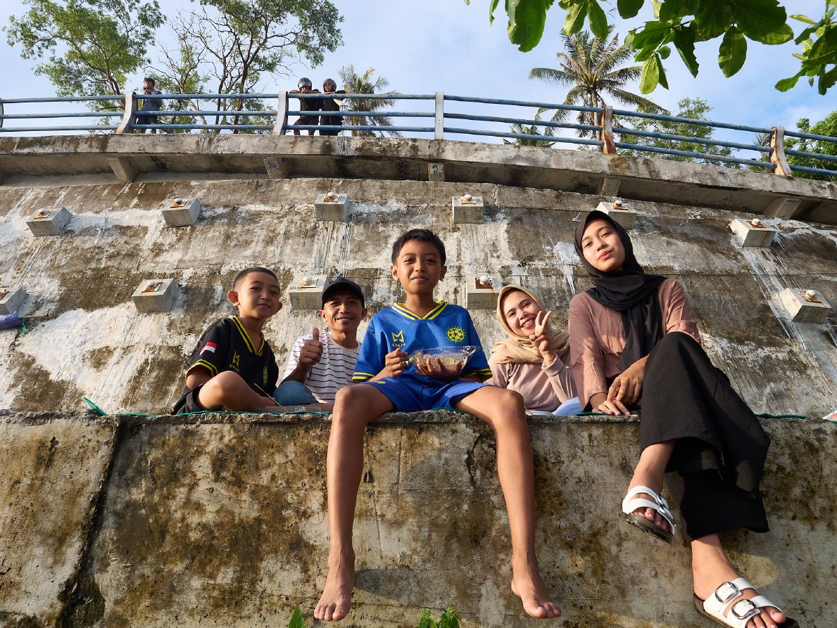 A family sits together on a weathered concrete wall, enjoying a sunny day with palm trees swaying in the background.