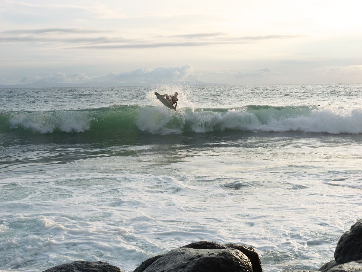 A surfer launches off the crest of a turquoise wave as the sun sets over the calm Lombok horizon.