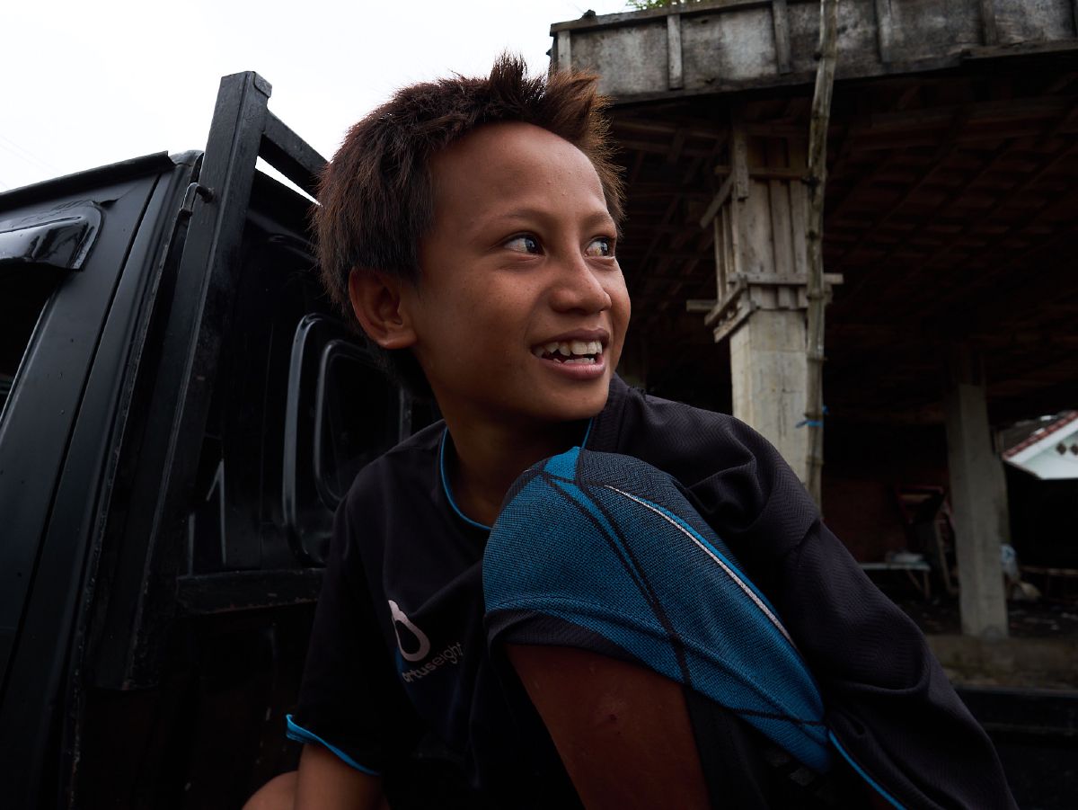 A young boy with a backpack smiles brightly while standing beside a vehicle in rural Lombok.