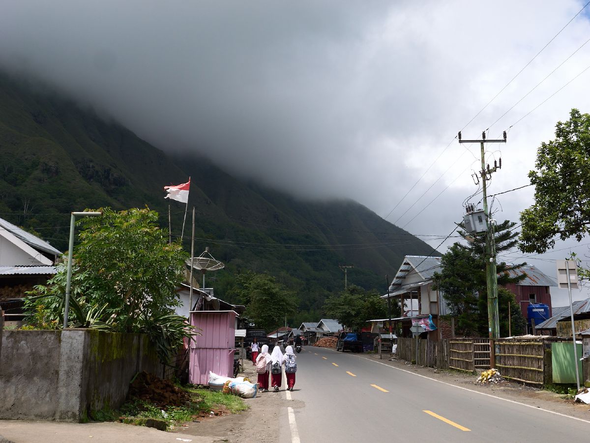 Three women in traditional dress walk along a village road with a misty mountain looming in the background under an Indonesian flag in Lombok.
