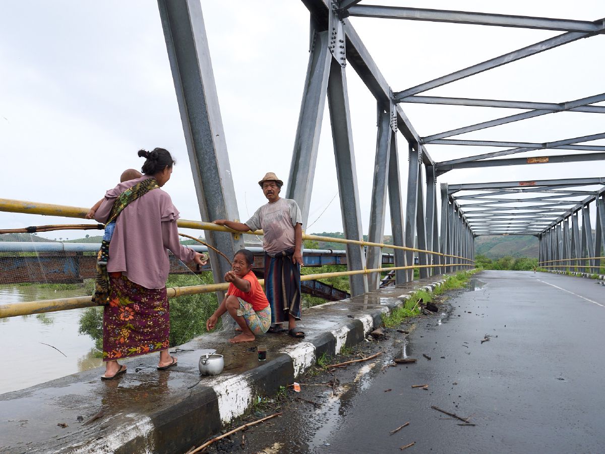 Three local residents conversing on a steel bridge beside a river in Lombok.