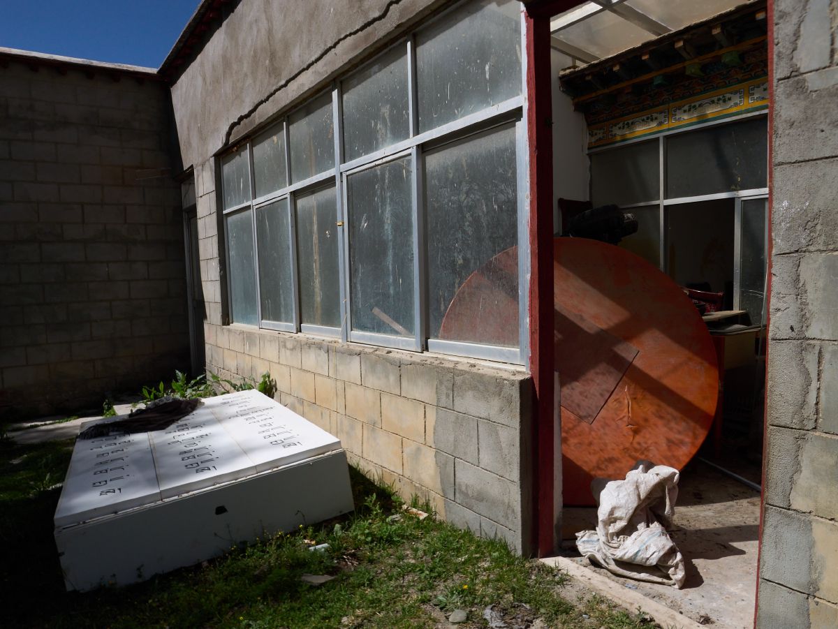 Sunlit corner of a rural building featuring a large wooden disc and discarded appliance in a grassy yard near Lhasa.