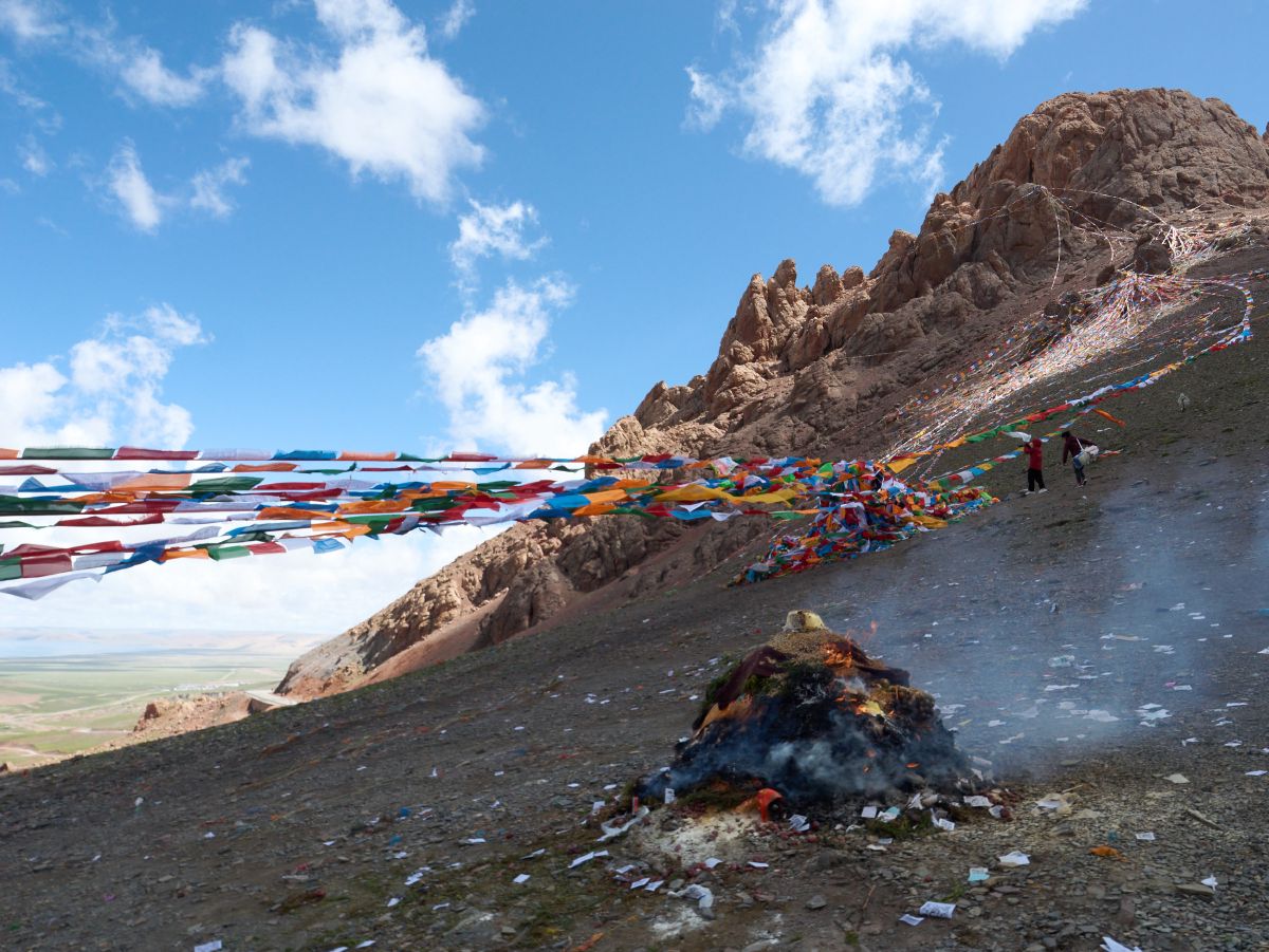 Colorful prayer flags fluttering across a mountain pass with a smoking ceremonial fire and rugged peaks near Lhasa.