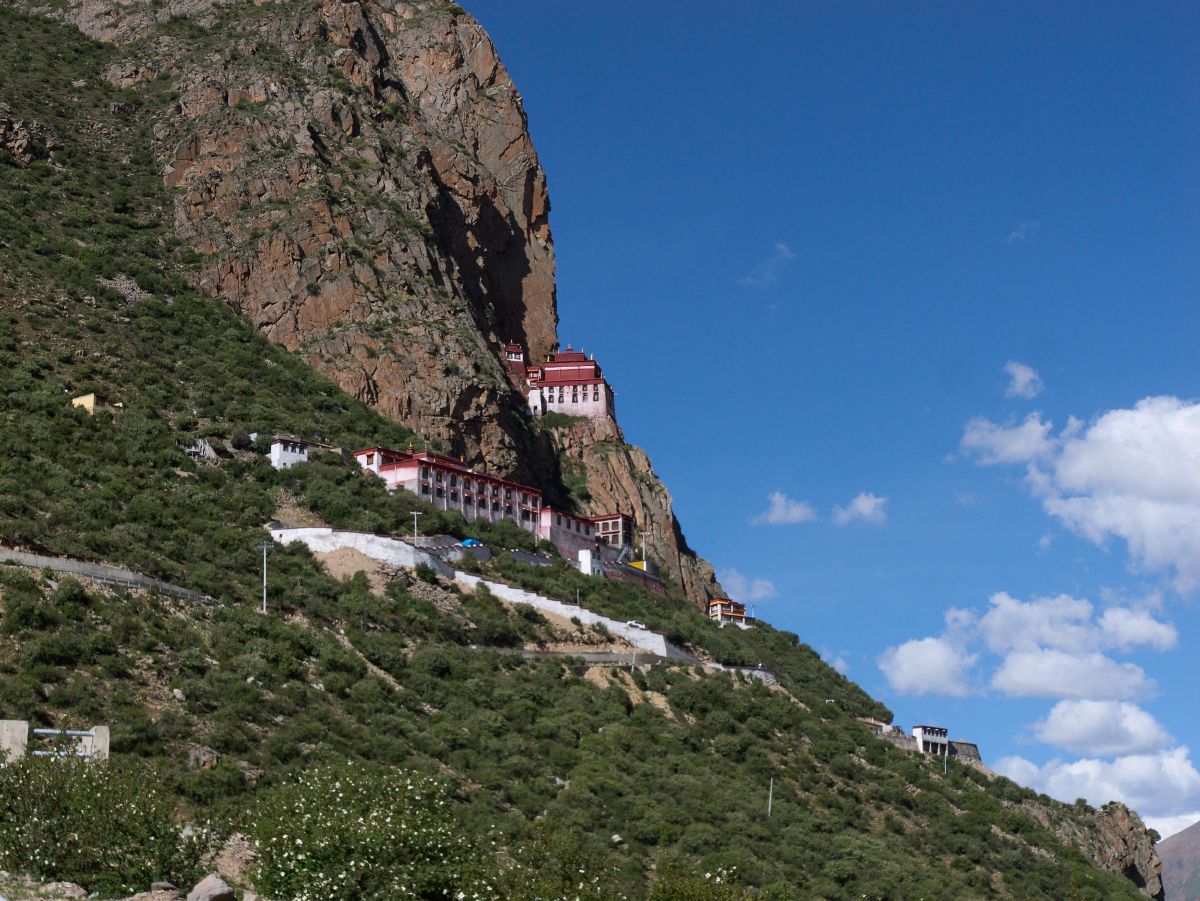 A traditional Tibetan monastery complex built into the side of a steep rocky cliff with red-roofed buildings and white walls cascading down the mountainside near Lhasa.