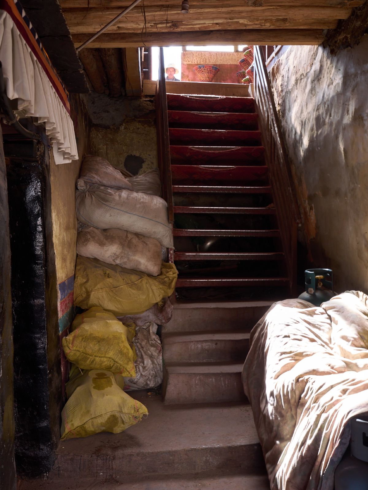 A narrow interior stairway lined with stacked sacks and bedding leads upward toward a brightly lit opening in a traditional building near Lhasa.