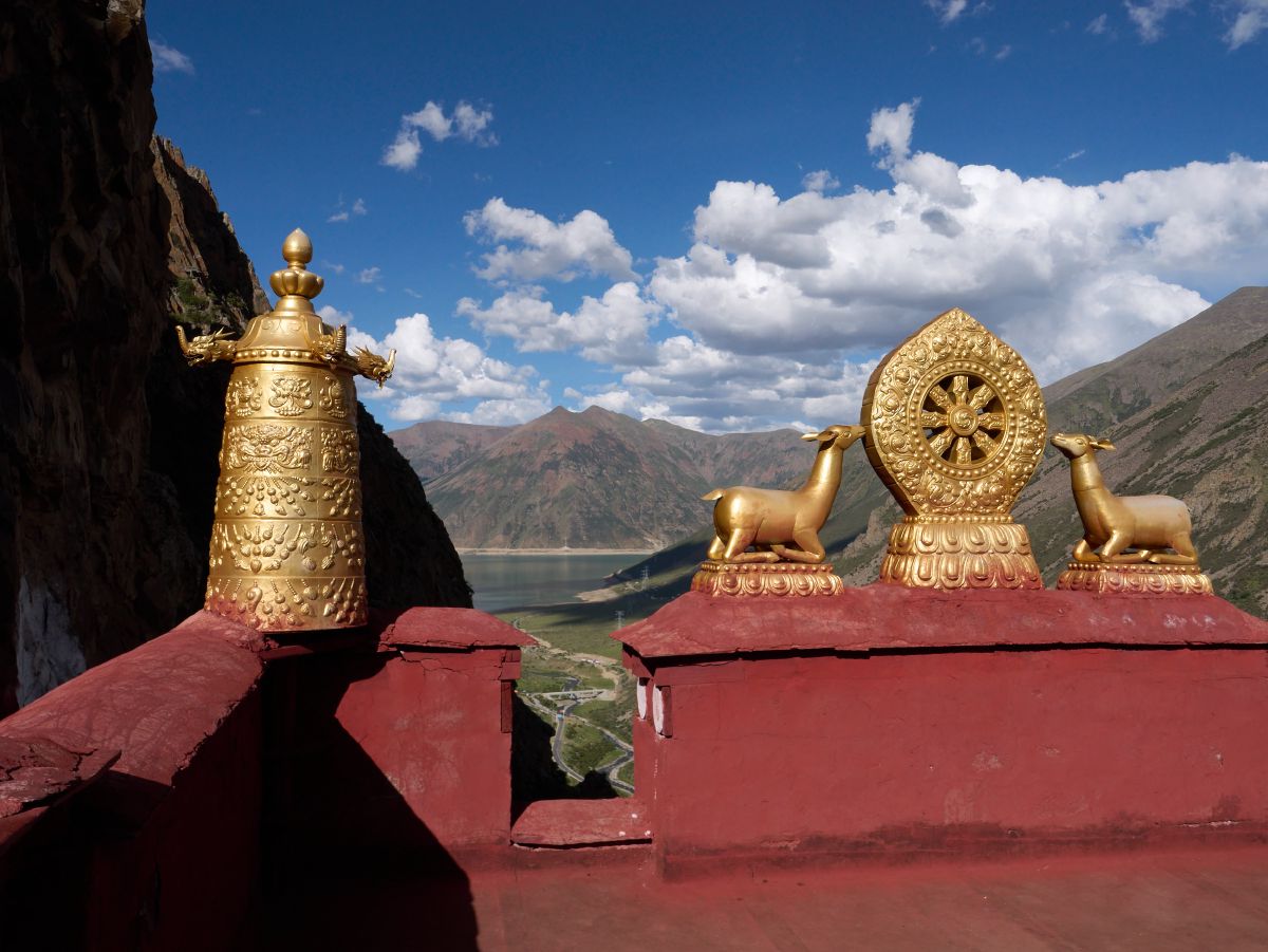 Ornate golden Buddhist statues including a dharma wheel flanked by deer sit atop red pedestals overlooking a vast mountain valley and lake near Lhasa.