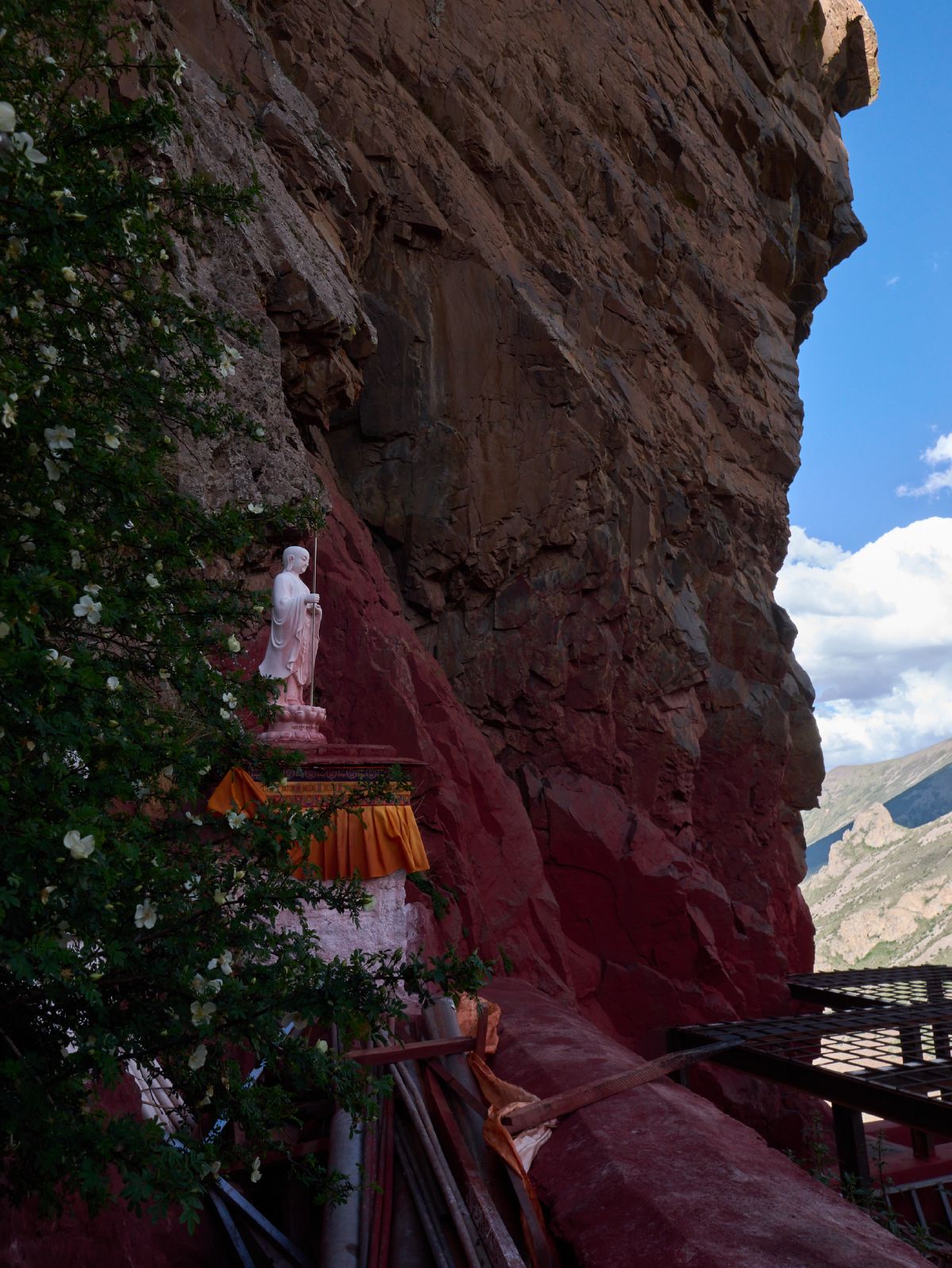 A white Buddha statue stands serenely on a red-painted cliffside platform, draped with golden fabric and surrounded by flowering shrubs.