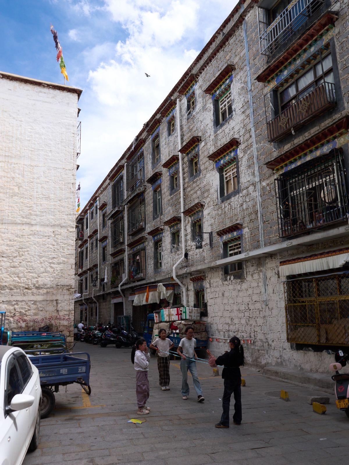Children play with a jump rope in a narrow street lined with traditional whitewashed Tibetan buildings under a bright blue sky.