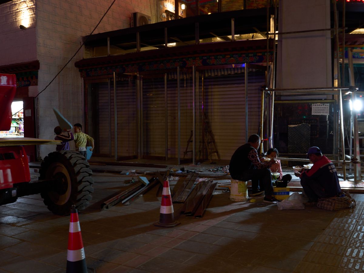 Construction workers take a break to eat dinner on the sidewalk at night, surrounded by scaffolding and traffic cones near a building under renovation.