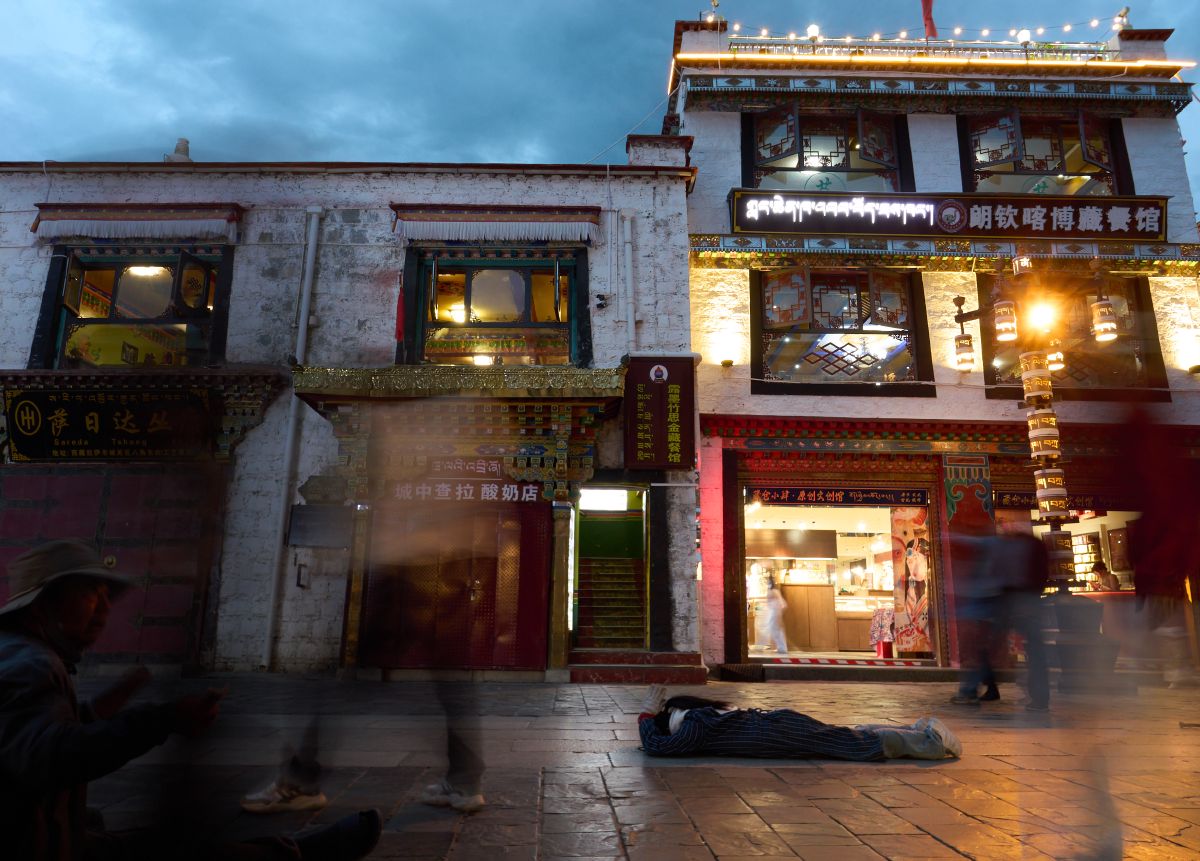 A pilgrim lies prostrate on the illuminated street at dusk, while blurred figures walk past traditional Tibetan buildings adorned with glowing signs.