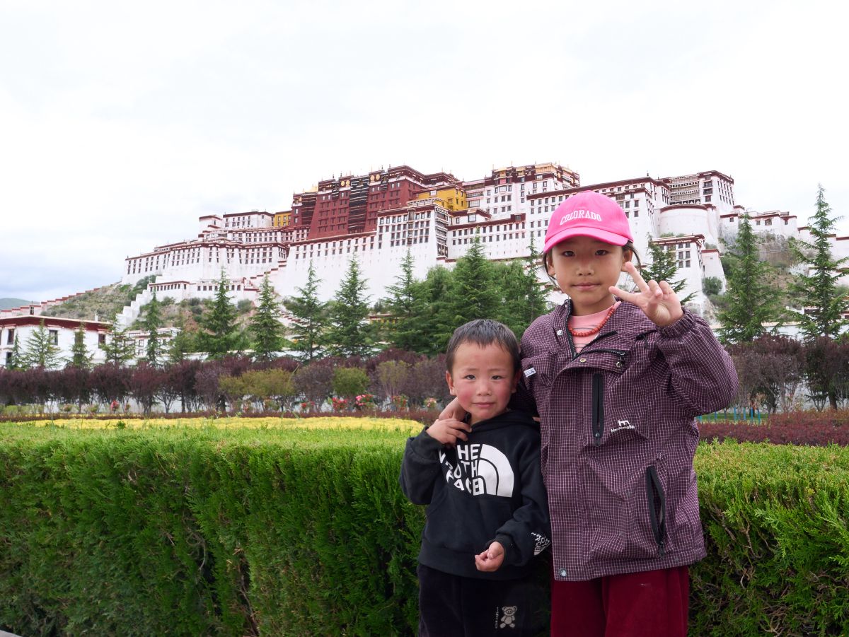 Two children pose happily in front of the iconic Potala Palace, with the younger sibling flashing a peace sign.